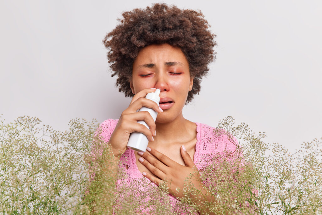 Unhappy curly haired woman suffers from runny nose uses nasal spray has red swelling eyes has allergic rhinitis reaction on wild plant poses against white background. People and sickness concept