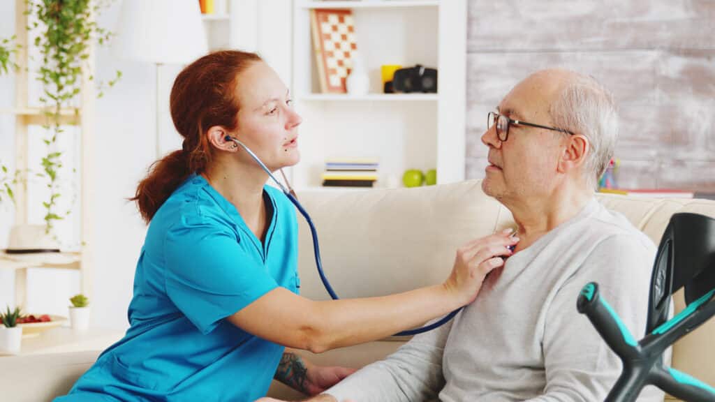 Caucasian female nurse listening to elderly retired man heartbeat in bright and cozy nursing home