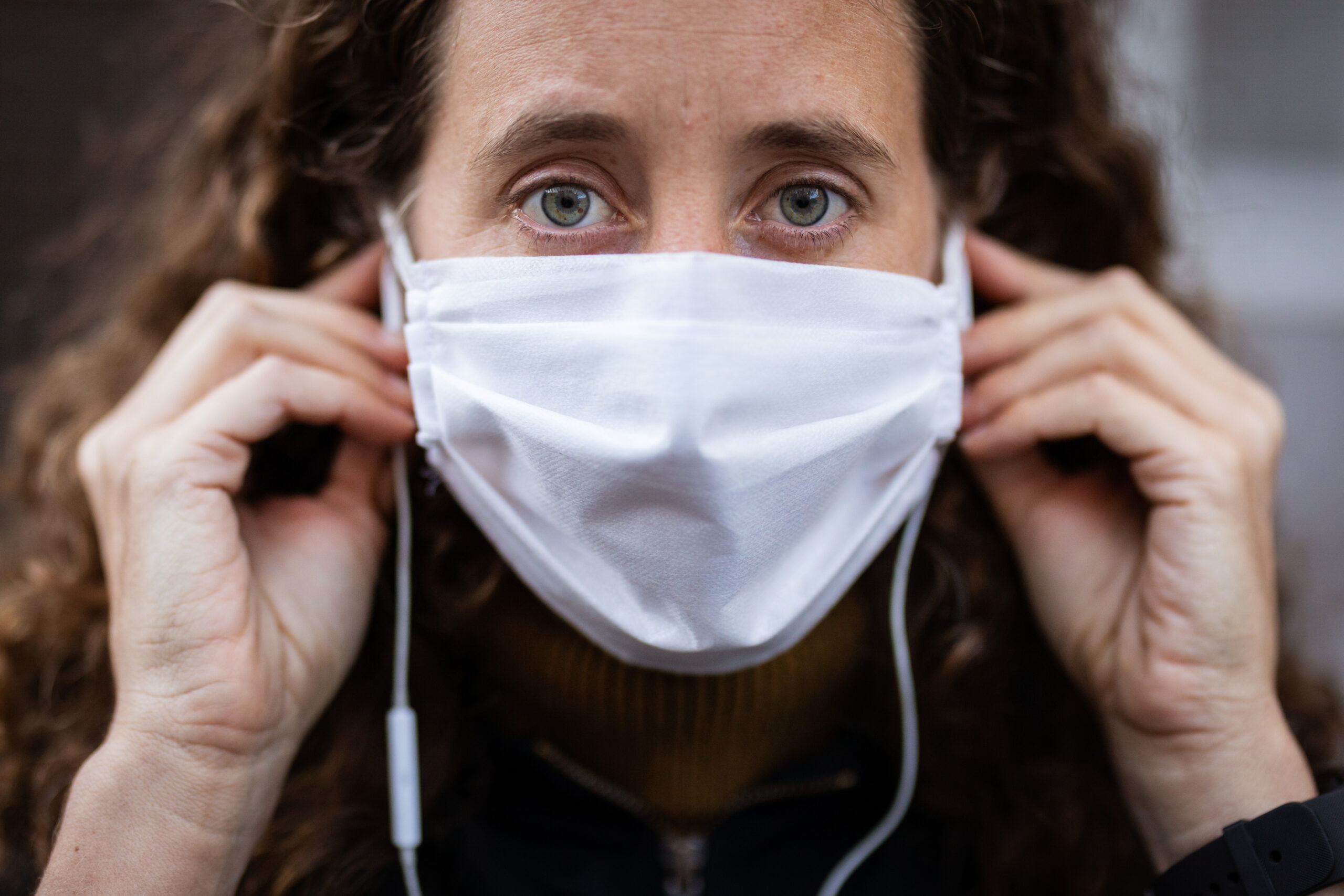 Portrait close up of Caucasian woman out and about in the city streets during the day, wearing a face mask against covid19 coronavirus, putting on earphones and looking straight to camera