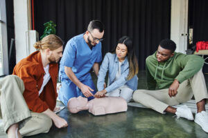 young asian woman practicing life saving skills by doing chest compressions on CPR manikin near multiethnic team and medical instructor, cardiopulmonary resuscitation, first aid training seminar adult CPR