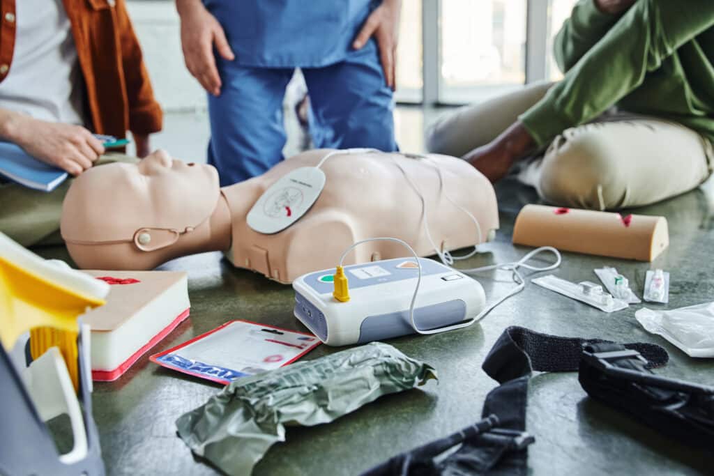 CPR manikin, defibrillator, wound care simulators, compression tourniquet, bandages and syringes near instructor and multiethnic participants of first aid seminar, emergency preparedness concept
