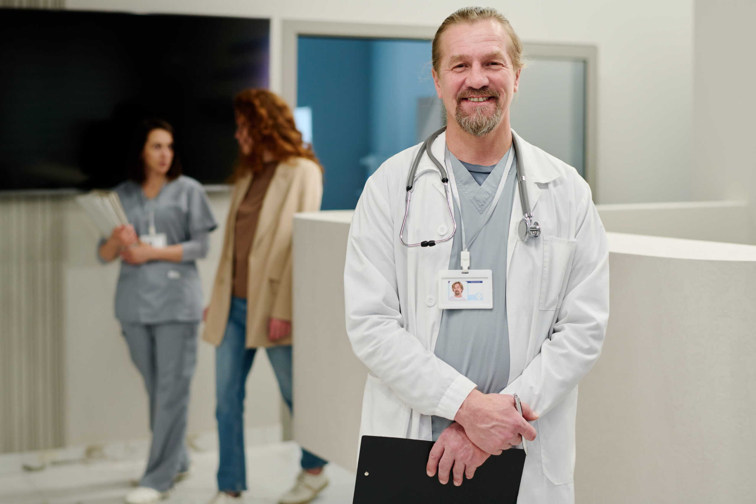 Successful male clinician in labcoat looking at camera with smile while standing against reception counter and two young women