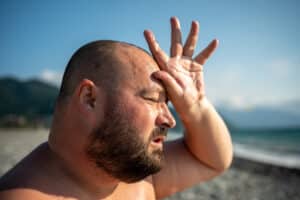 Overweight man suffers heatstroke on beach, shields head with hand. Struggles with heat, wipes sweat, squints under sun, challenges of excess weight, summer vacation discomfort, weather risks