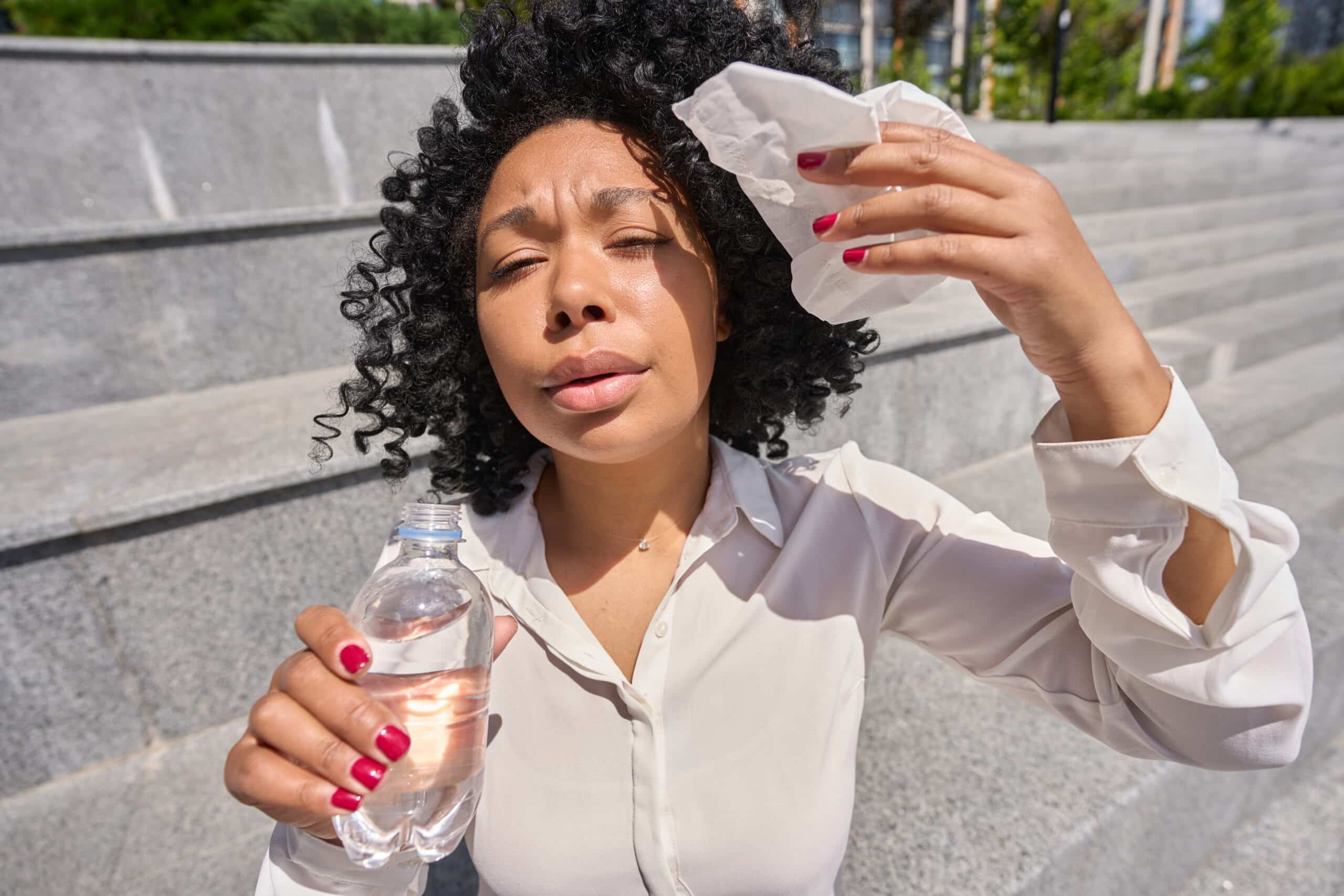Multiracial woman sitting in the heatstroke wiping herself with a napkin and drinking water