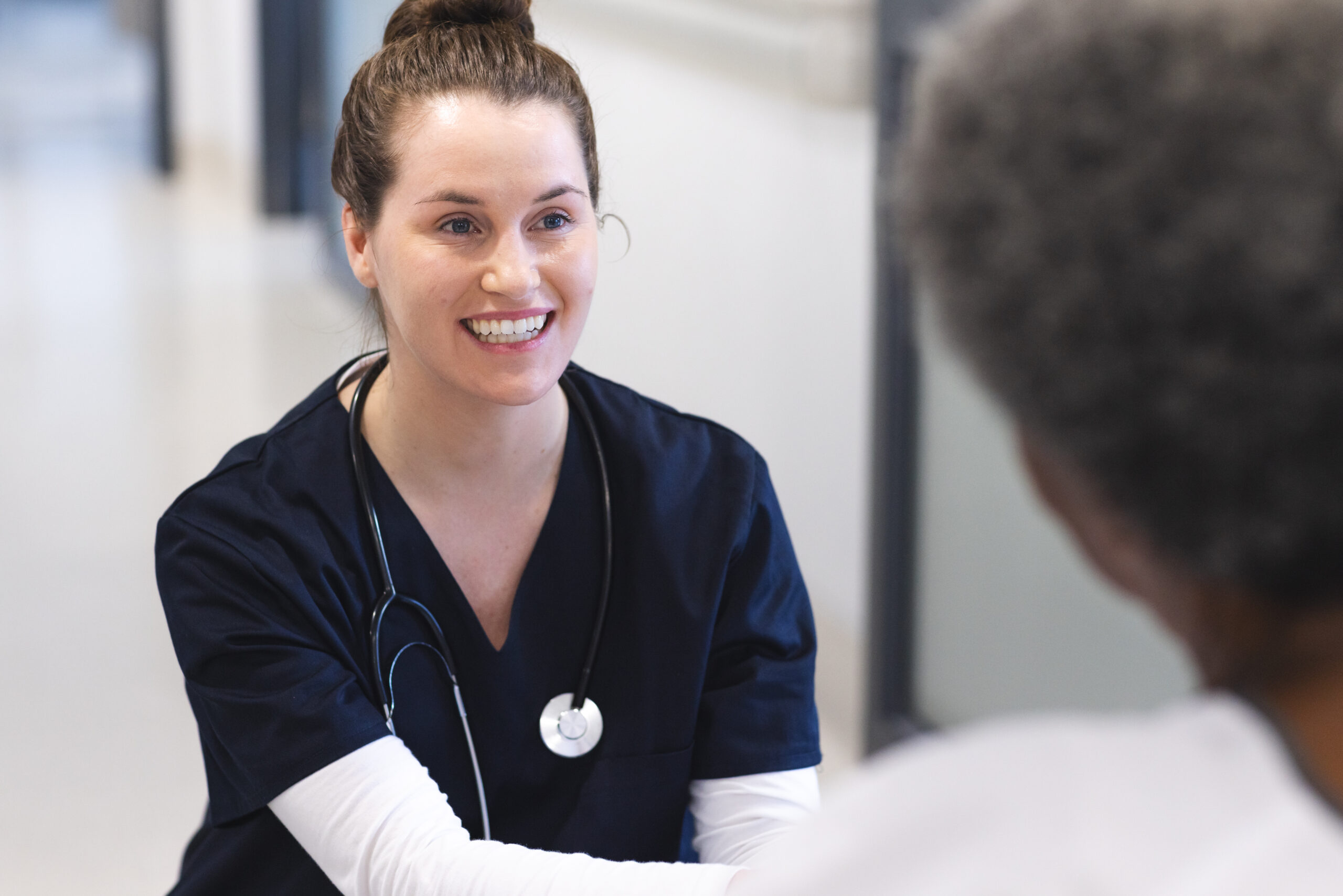 Happy diverse female doctor talking with senior female patient in wheelchair in hospital corridor. Medicine, healthcare and medical services, unaltered.
