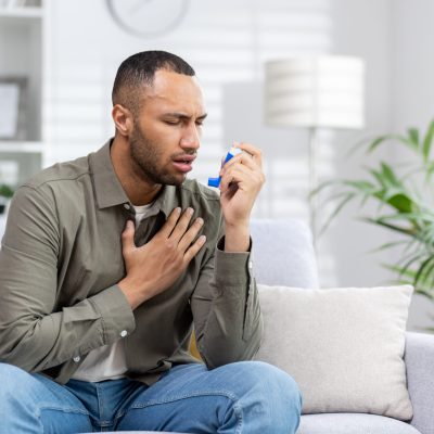 A young American man is sitting on the couch at home and having an asthma attack. Holds his chest and breathes hard, blows an inhaler into his mouth