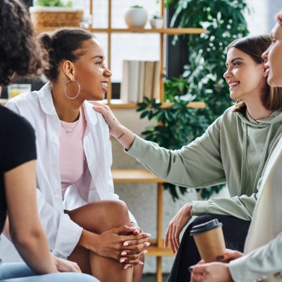 young woman touching shoulder of pleased african american girlfriend while sitting next to redhead psychologist with paper cup during therapy in consulting room, mental wellness concept